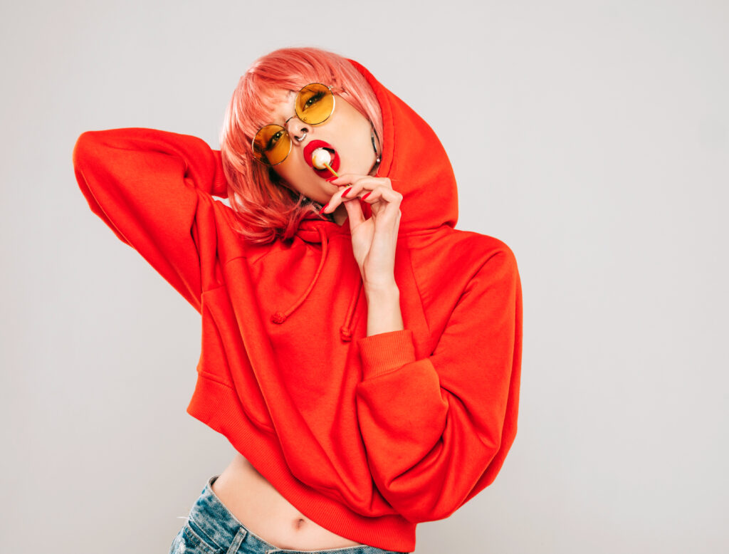 Młoda piękna hipsterska zła dziewczyna w modnej czerwonej letniej czerwonej bluzie z kapturem i kolczyku w nosie.Seksowna beztroska kobieta pozująca w studio na szarym tle w peruce.Gorąca modelka liże okrągłe cukierki